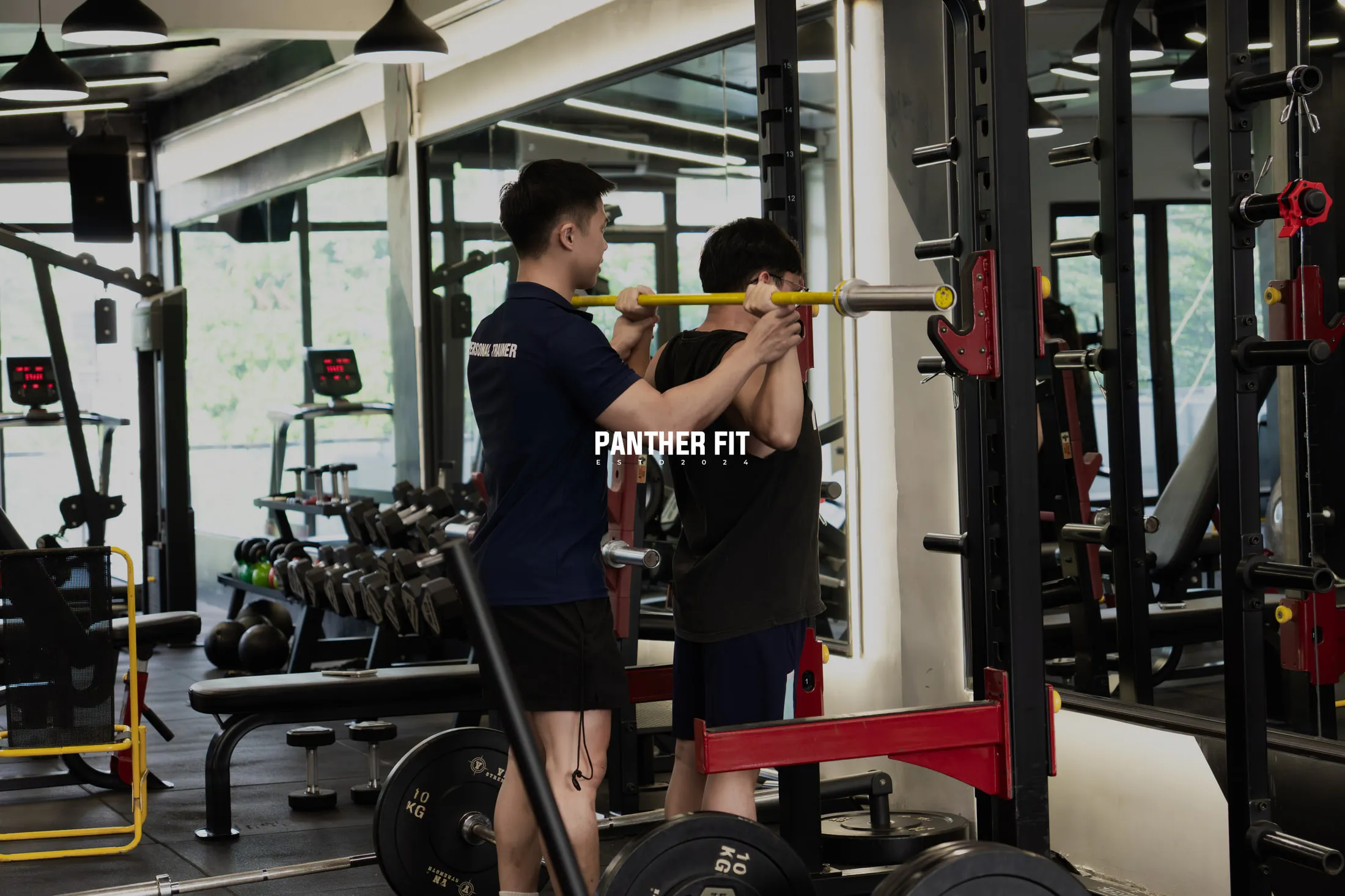 Huấn luyện viên Panther Fit đứng cạnh hướng dẫn một hội viên nâng tạ đòn