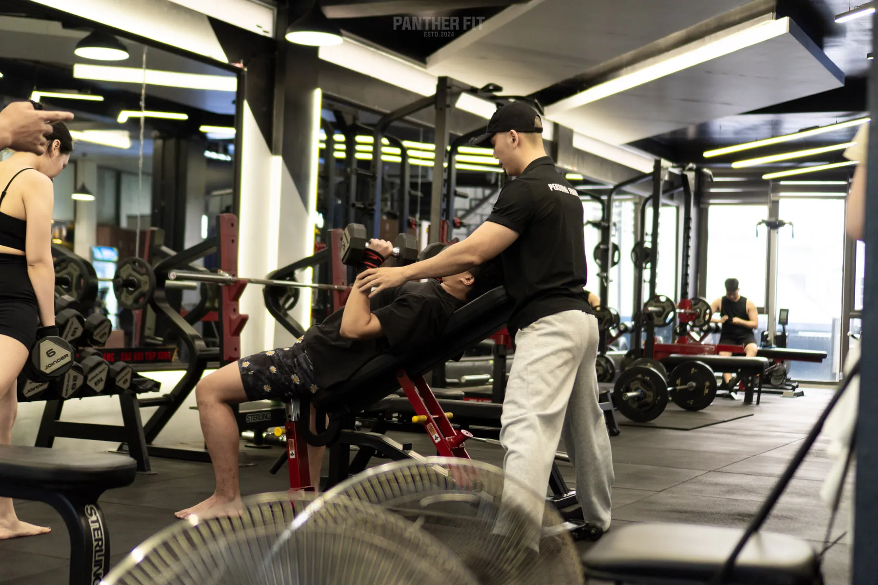 Huấn luyện viên đứng cạnh hỗ trợ hội viên đẩy tạ tay tại Panther Fit
