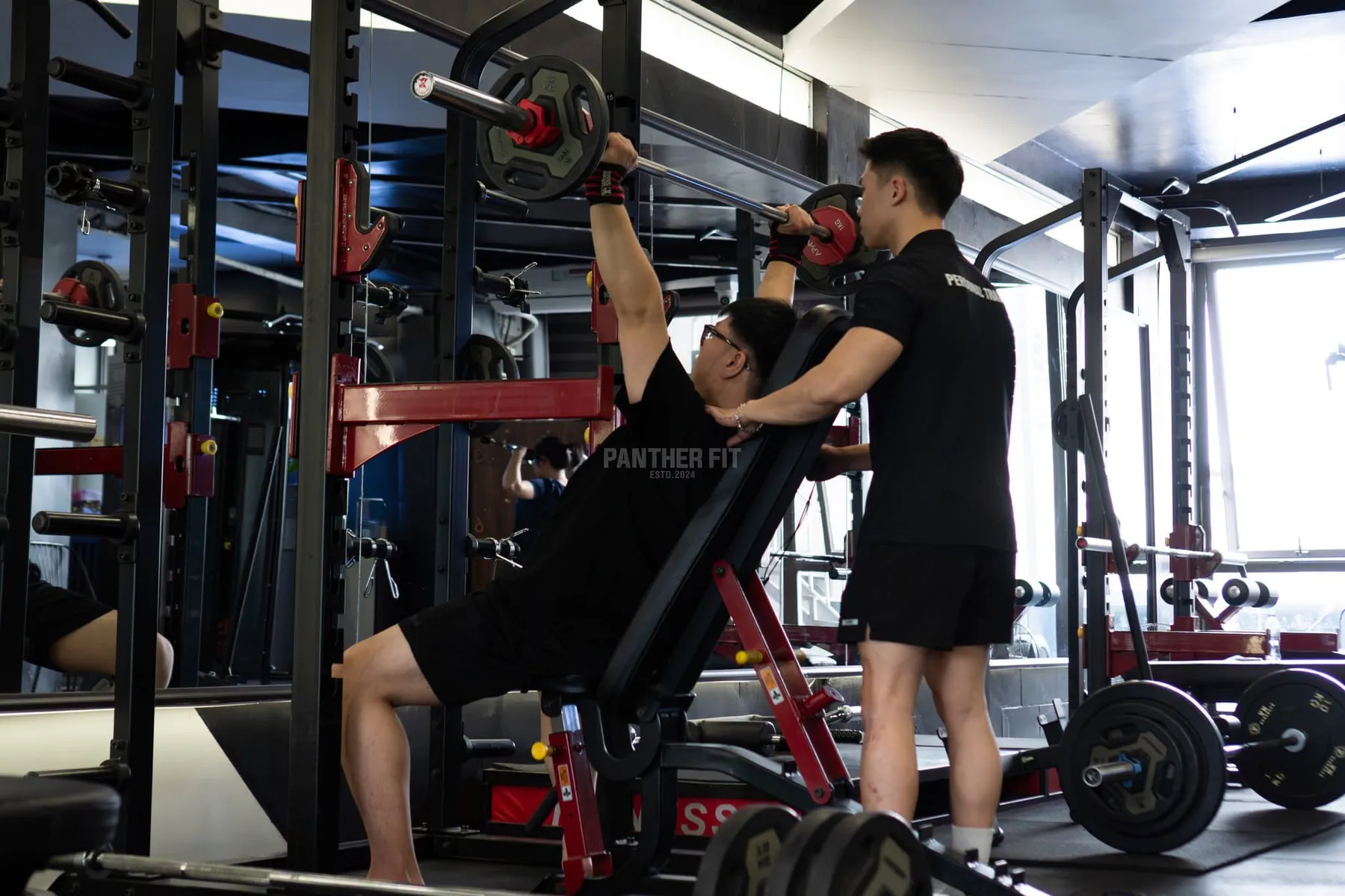 Huấn luyện viên đứng cạnh đỡ tạ cho hội viên đang đẩy ngực tại Panther Fit